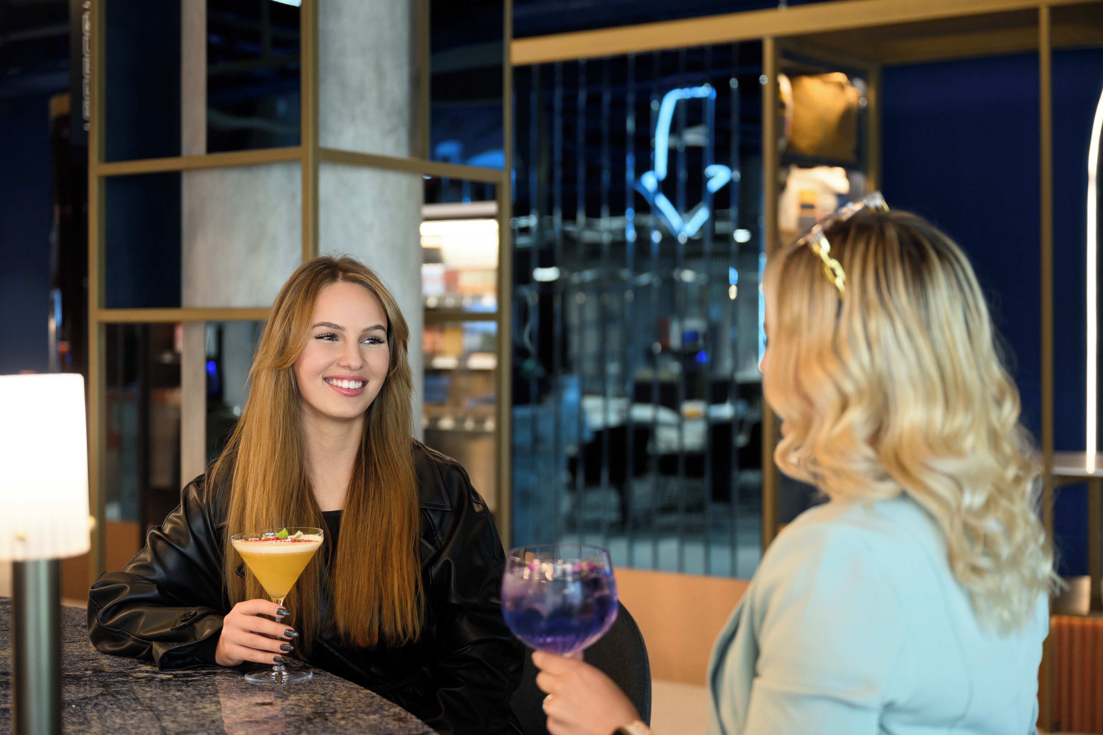 Two people sitting at a table with drinks, in a modern indoor setting with glass partitions and a neon sign.