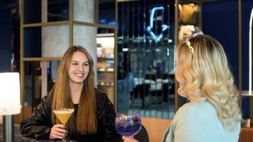 Two people sitting at a table with drinks, in a modern indoor setting with glass partitions and a neon sign.