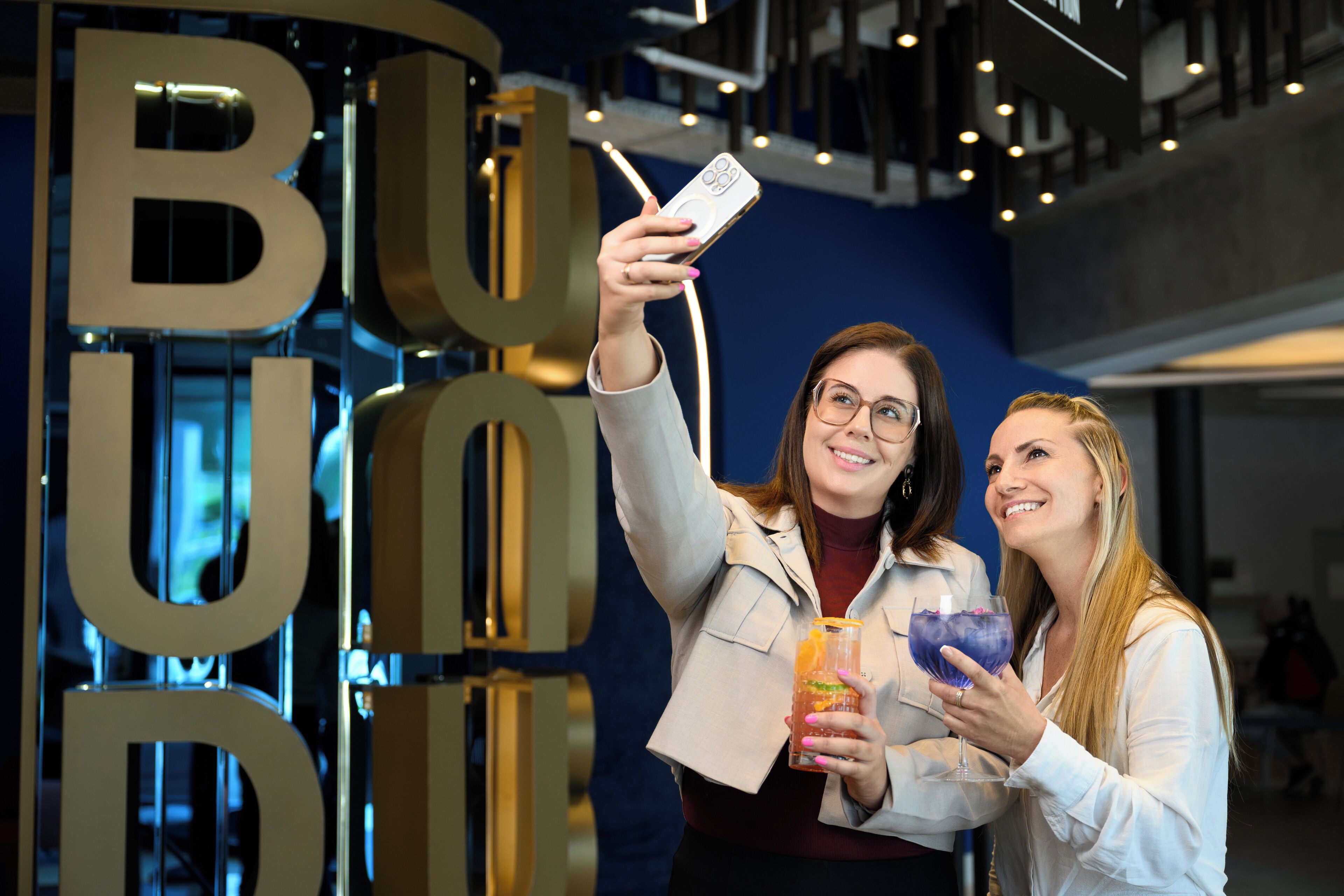 Dos personas tomándose un selfie con un cartel grande de 'BUD' al fondo.