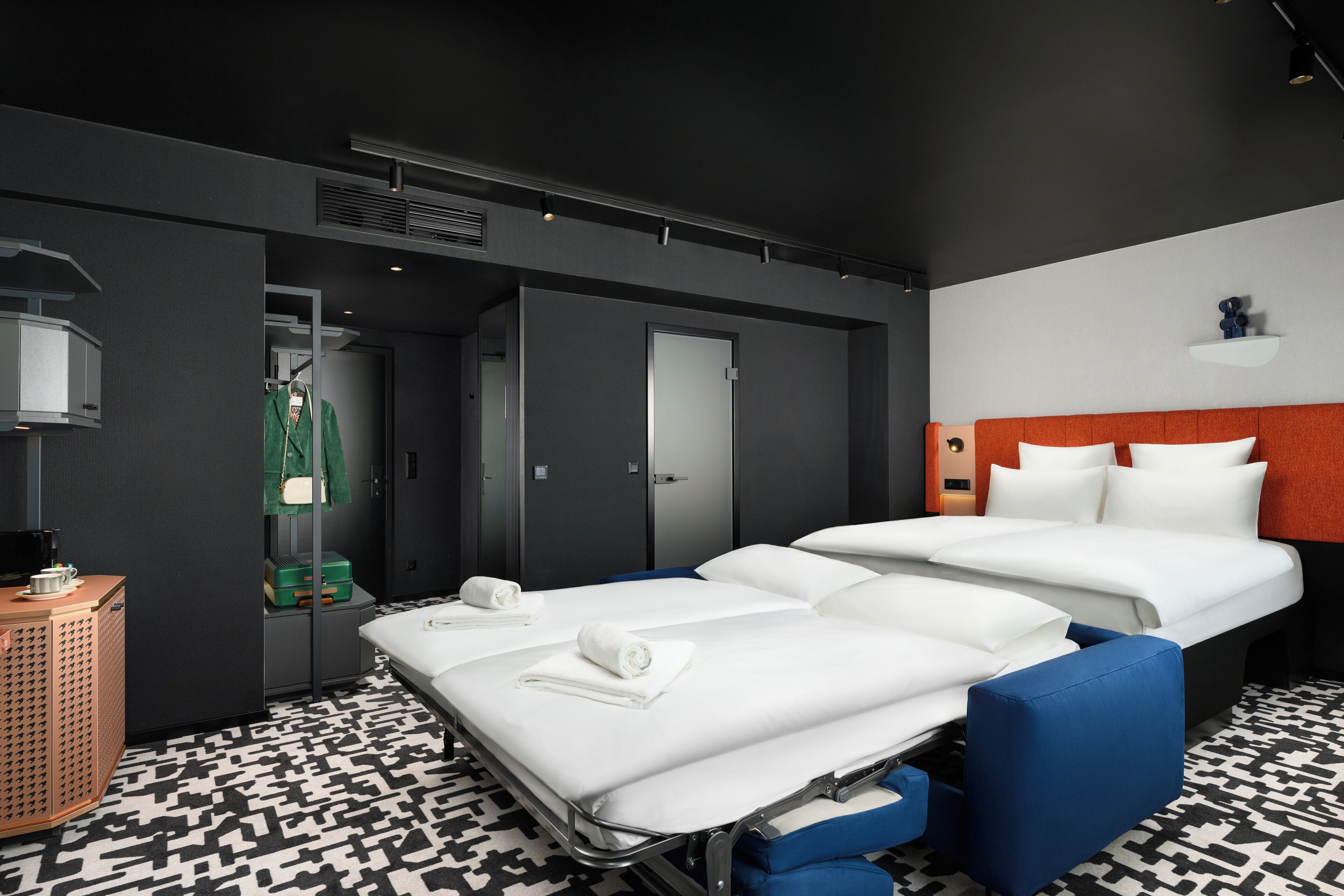 A hotel room with two beds, a small table with a lamp, a wardrobe, and a patterned floor.