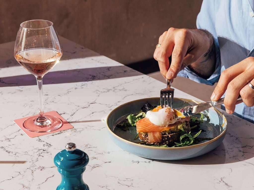Una persona cenando en una mesa con una copa de vino y un plato de comida.
