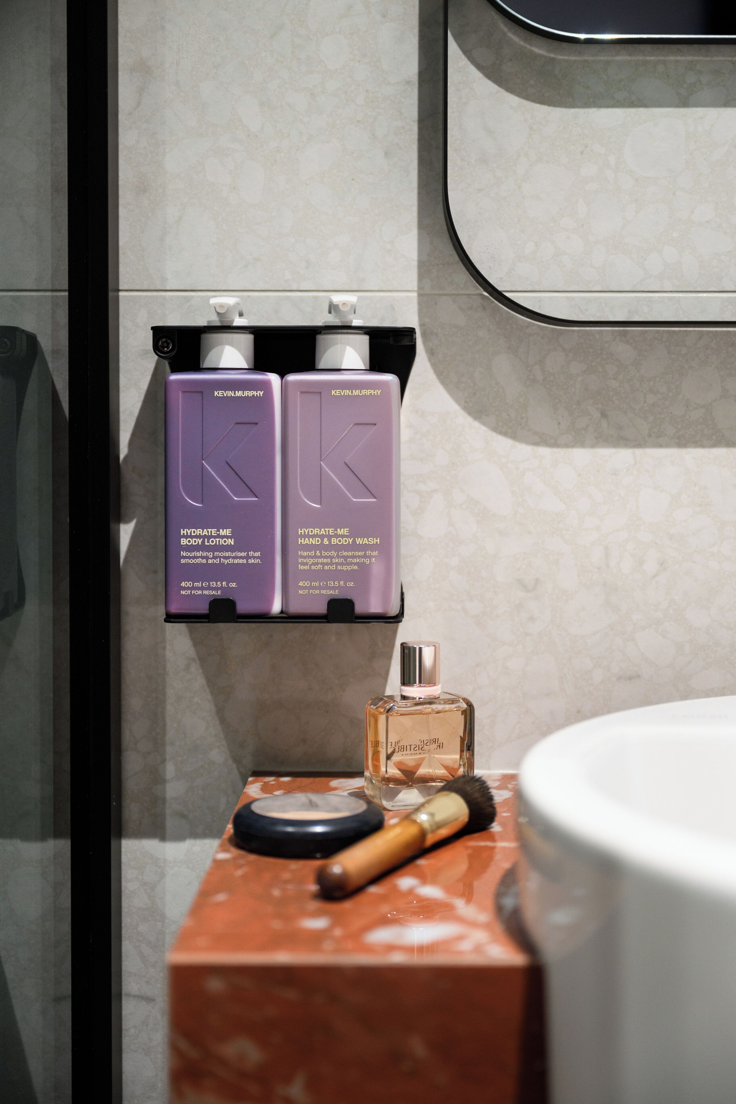 Um banheiro com bancada de mármore, dispensador de sabonete líquido, um frasco de perfume e duas embalagens de loção corporal da Kevin Murphy.