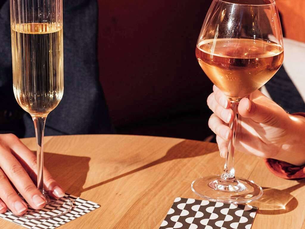 Dos personas sosteniendo copas de vino sobre una mesa de madera con posavasos decorados.