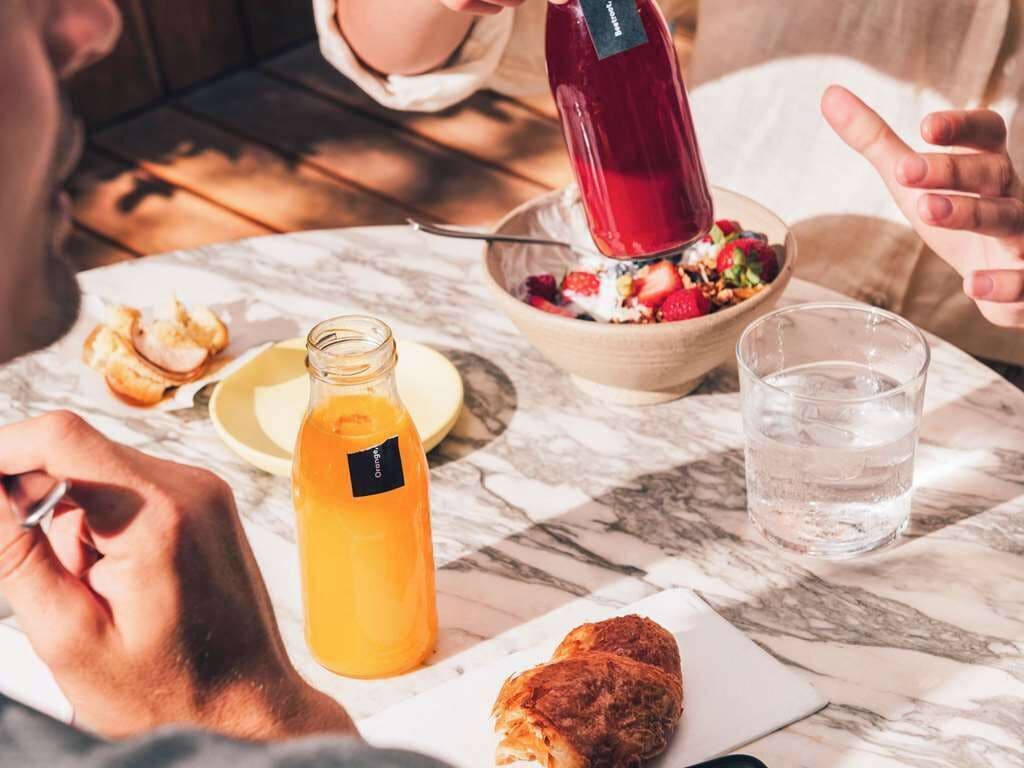 Una mesa de comedor con un tazón de ensalada, un vaso de agua, una botella de jugo de naranja y un pastel.