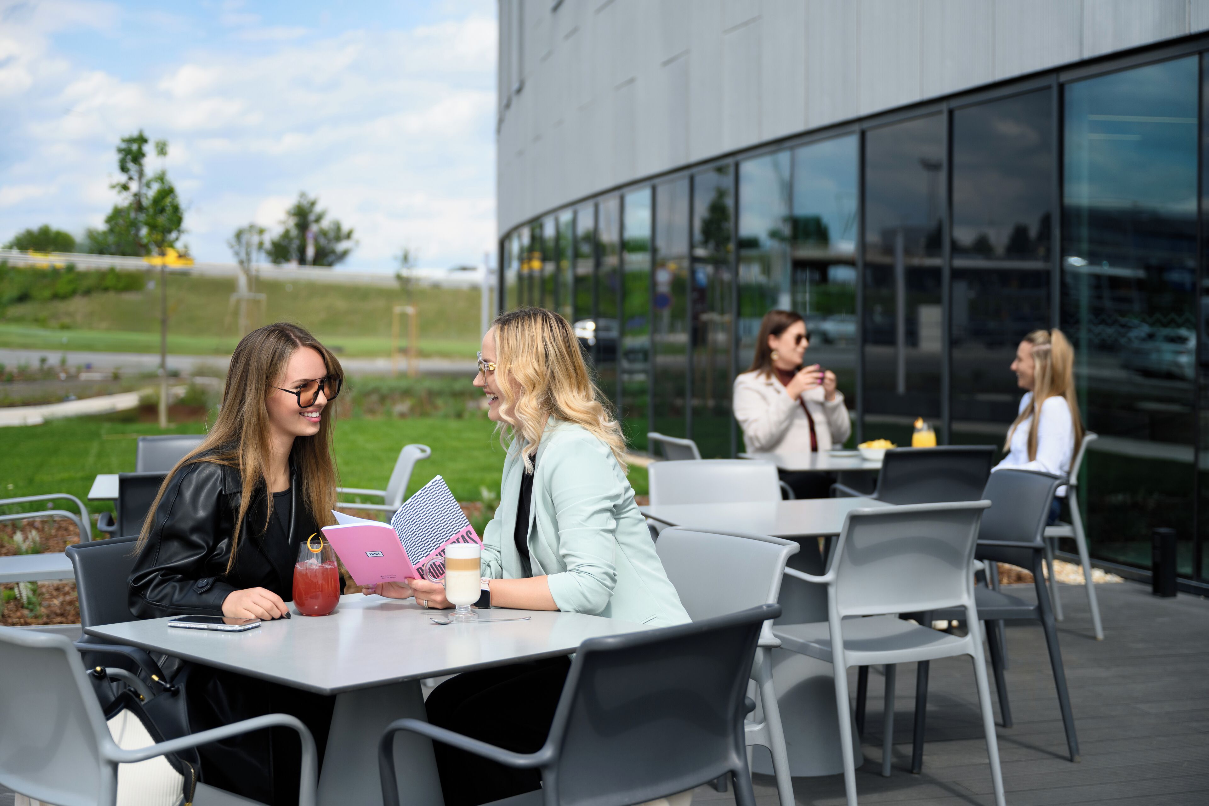 Ein Straßen-Café mit Tischen und Stühlen, ein Gebäude mit großen Fenstern und eine grünen Fläche im Hintergrund.