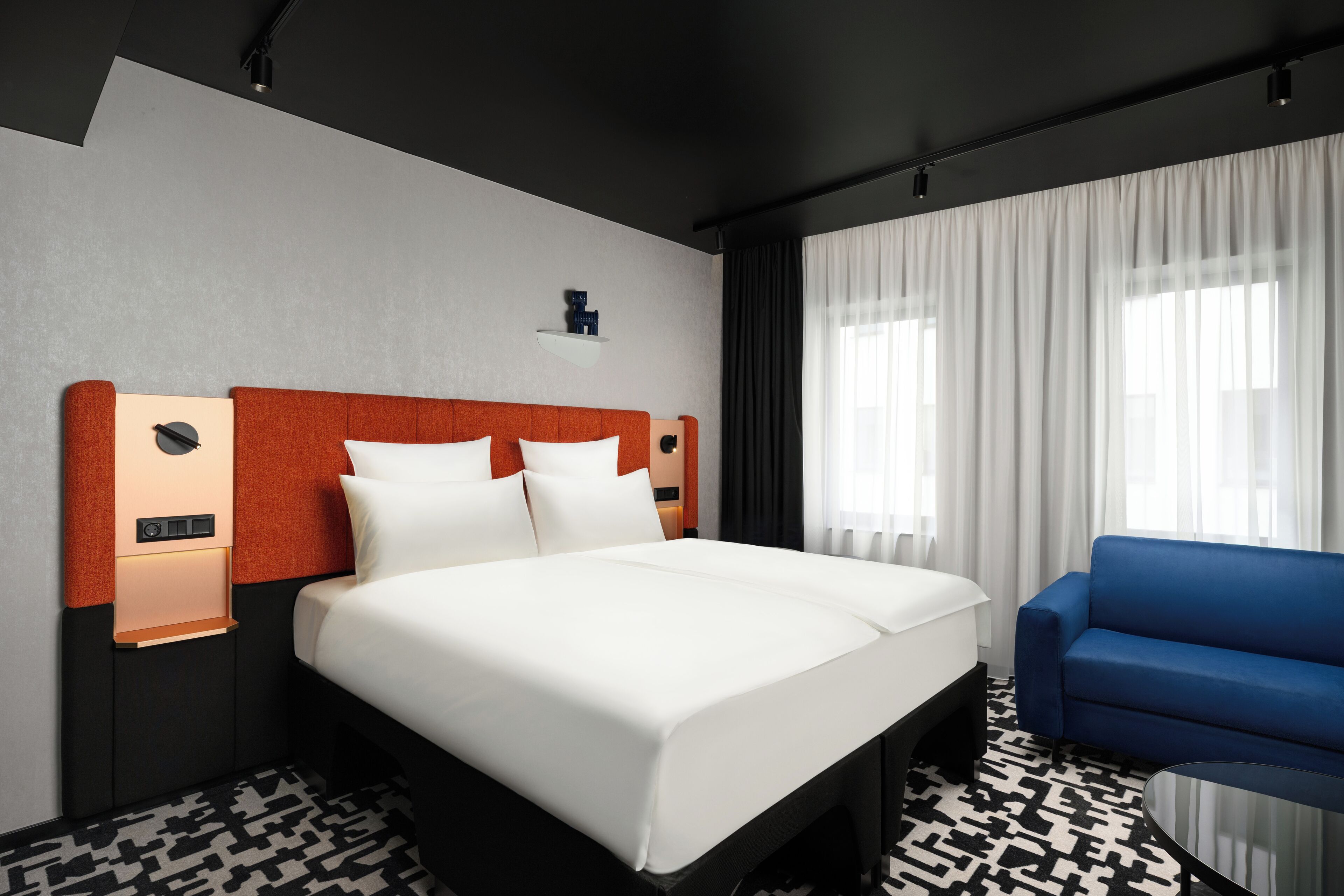 Habitación de hotel con una cama grande, un sillón azul, un estante en la pared y una ventana con cortinas.