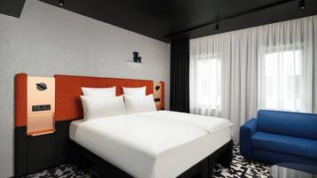 Habitación de hotel con una cama grande, un sillón azul, un estante en la pared y una ventana con cortinas.