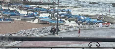 A coastal scene with boats docked along the shore, a pedestrian walkway, and a metal railing in the foreground.
