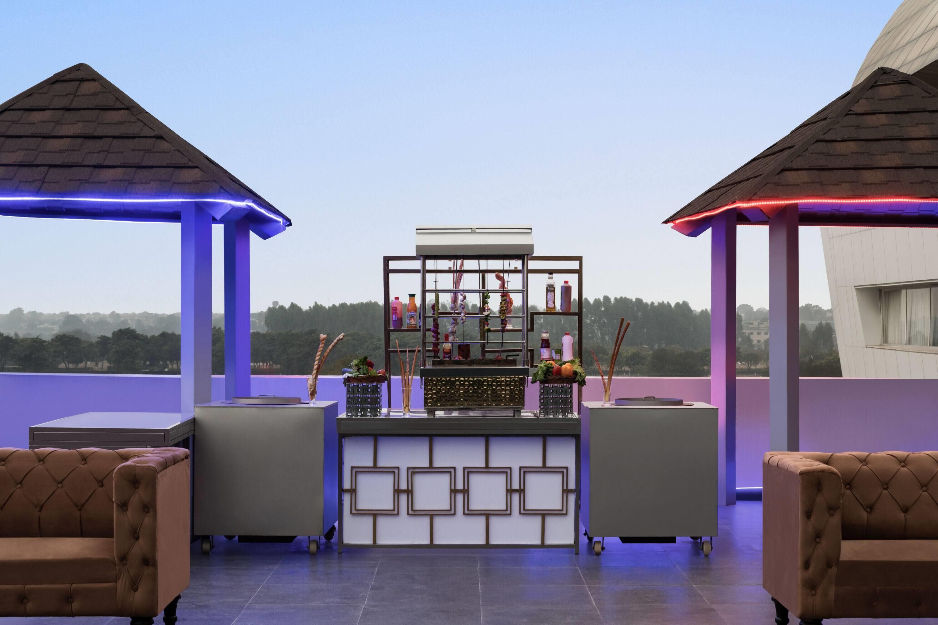 A rooftop bar with a bar counter, cushioned seating, and a view of the landscape.