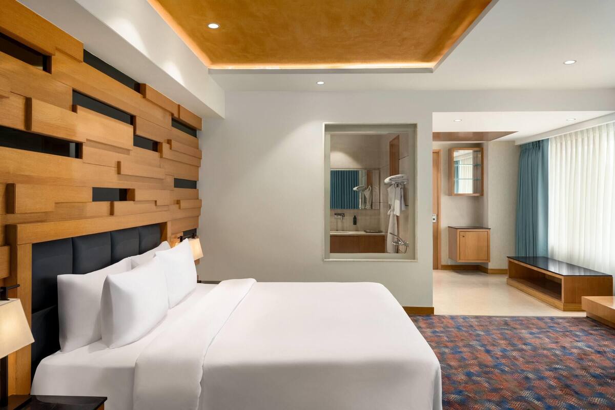 A modern hotel room with a large bed, a wooden headboard, a mirror, a bathroom visible through an open door, and a patterned carpet.