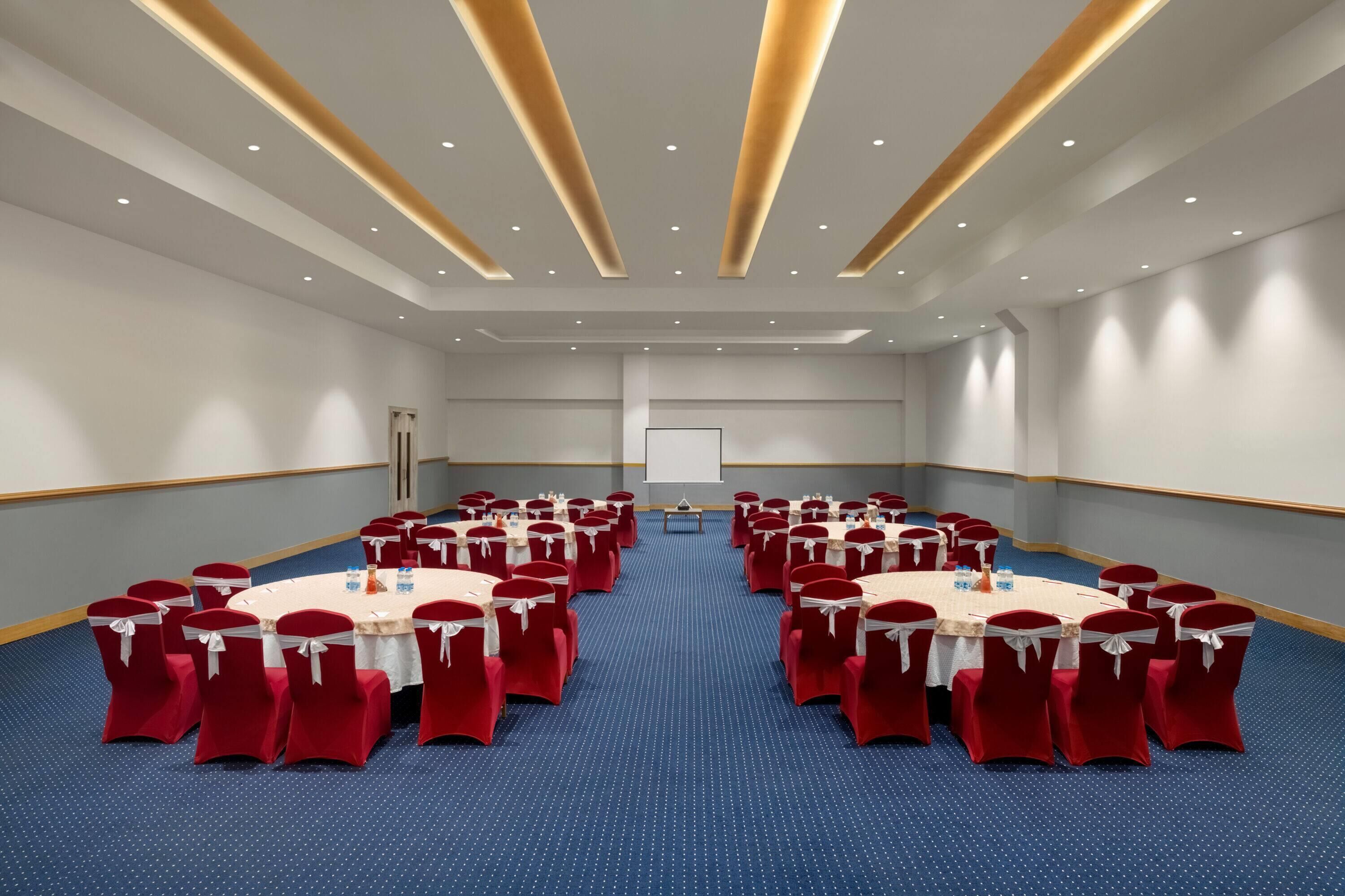 A conference room with round tables and red chairs, a projector screen, and a carpeted floor.