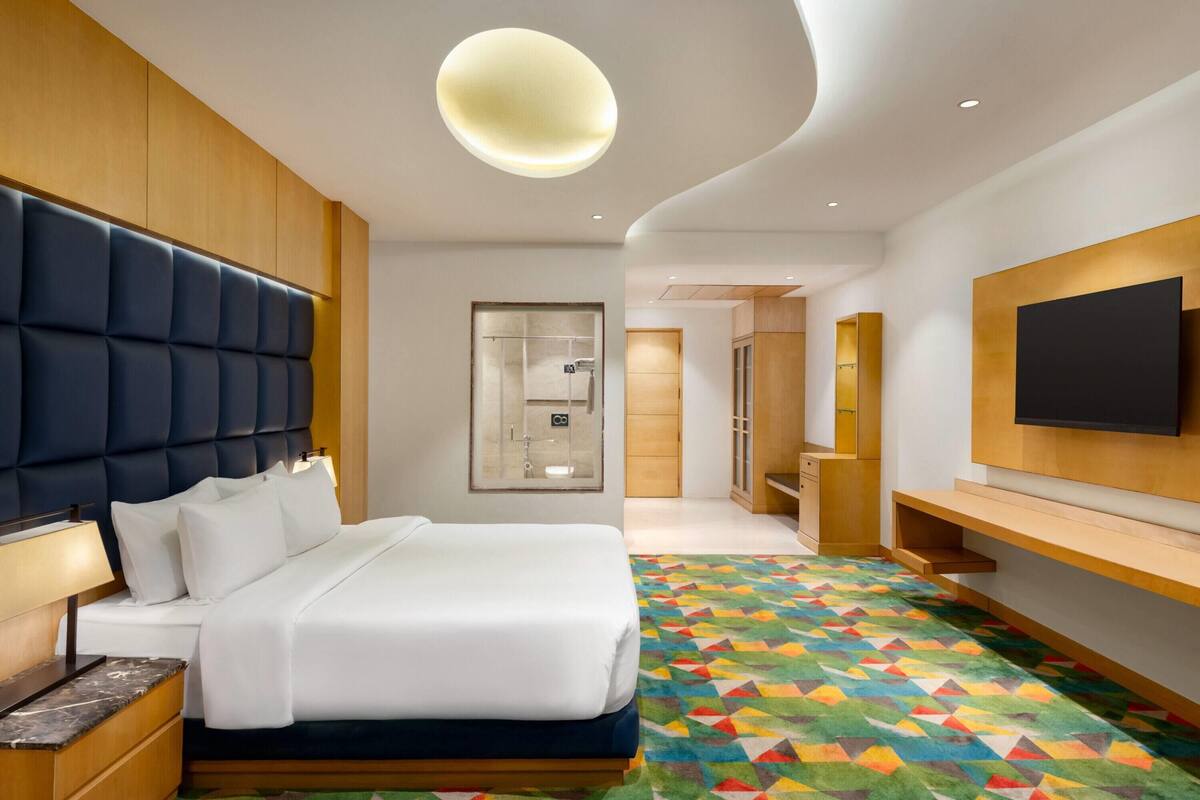 A modern hotel room with a large bed, a TV mounted on the wall, and a colorful geometric-patterned carpet.