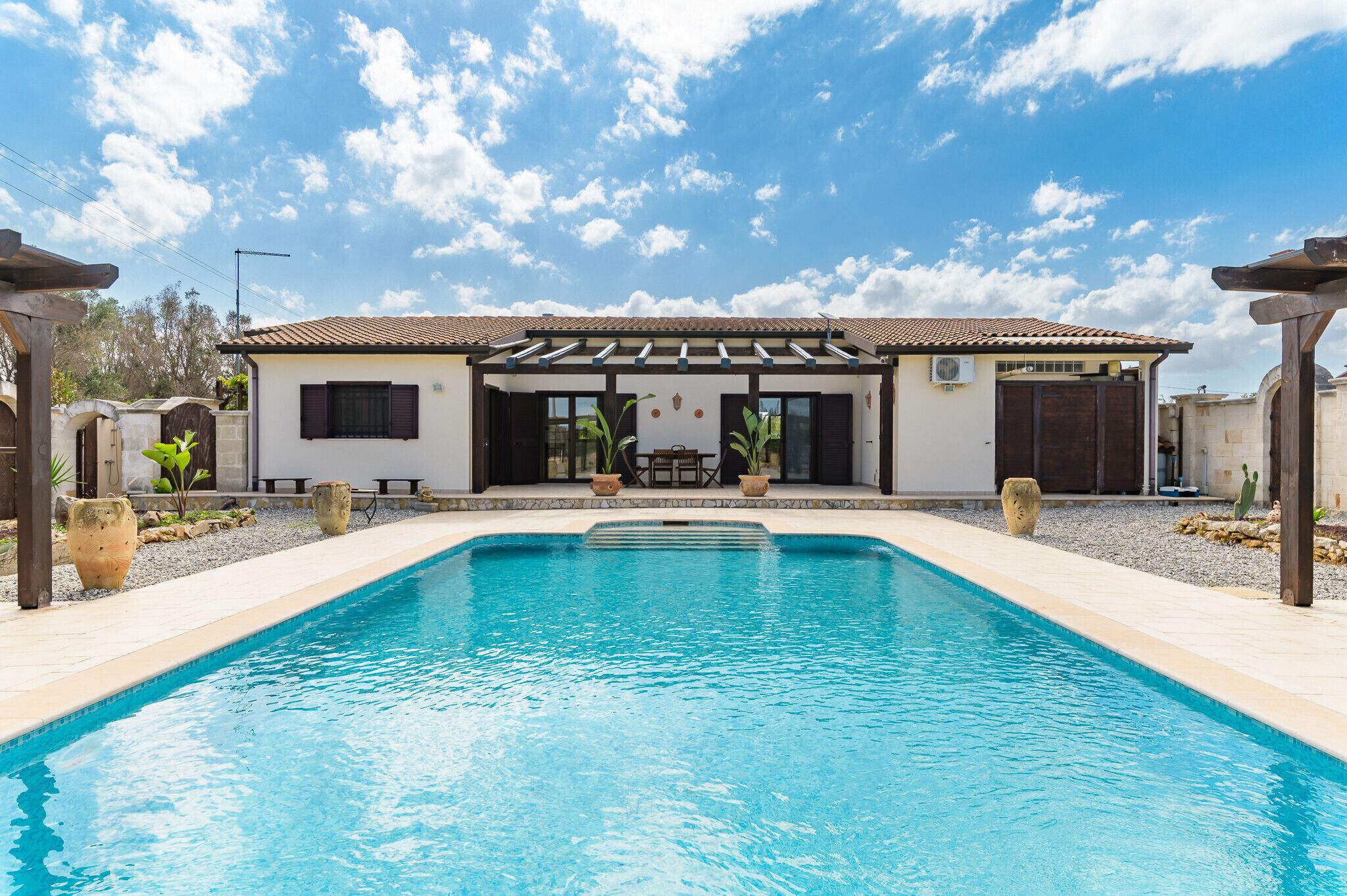 Ein Swimmingpool mit einer Pergola, einer überdachten Terrasse und Blick auf ein Gebäude mit einem Ziegeldach.