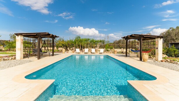Una piscina rectangular con agua azul cristalina, rodeada por una solana de baldosas. Hay dos pérgolas de madera con zonas de estar y una hamaca.