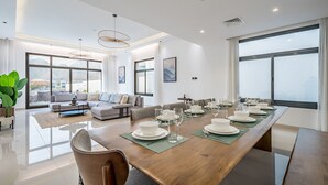 A spacious living room with a large dining table set for a meal, a sofa, and a large window with a mountain view. - Blue Marble Beach 4BR Villa with AsstRm,Private Pool and Access to Floating Pier (Fujairah)