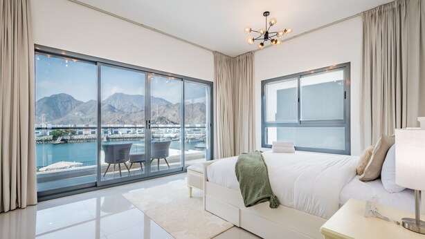 A modern hotel room with a large window offering a view of a marina and mountains. - Blue Marble Beach 4BR Villa with AsstRm,Private Pool and Access to Floating Pier (Fujairah)