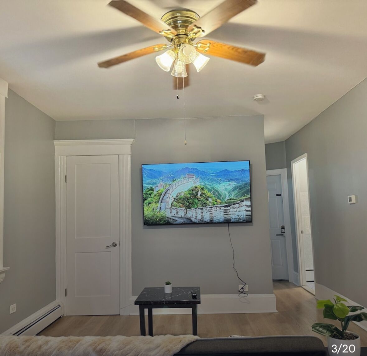 A living room with a ceiling fan, a mounted TV displaying a scenic image, a small table with a plant, and a door leading to another room.