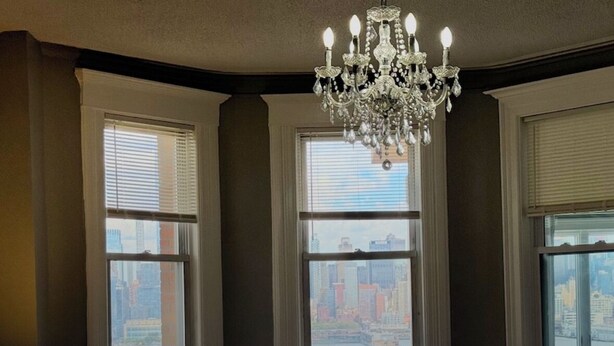A bedroom with a bed, a chandelier, and three windows with city views.