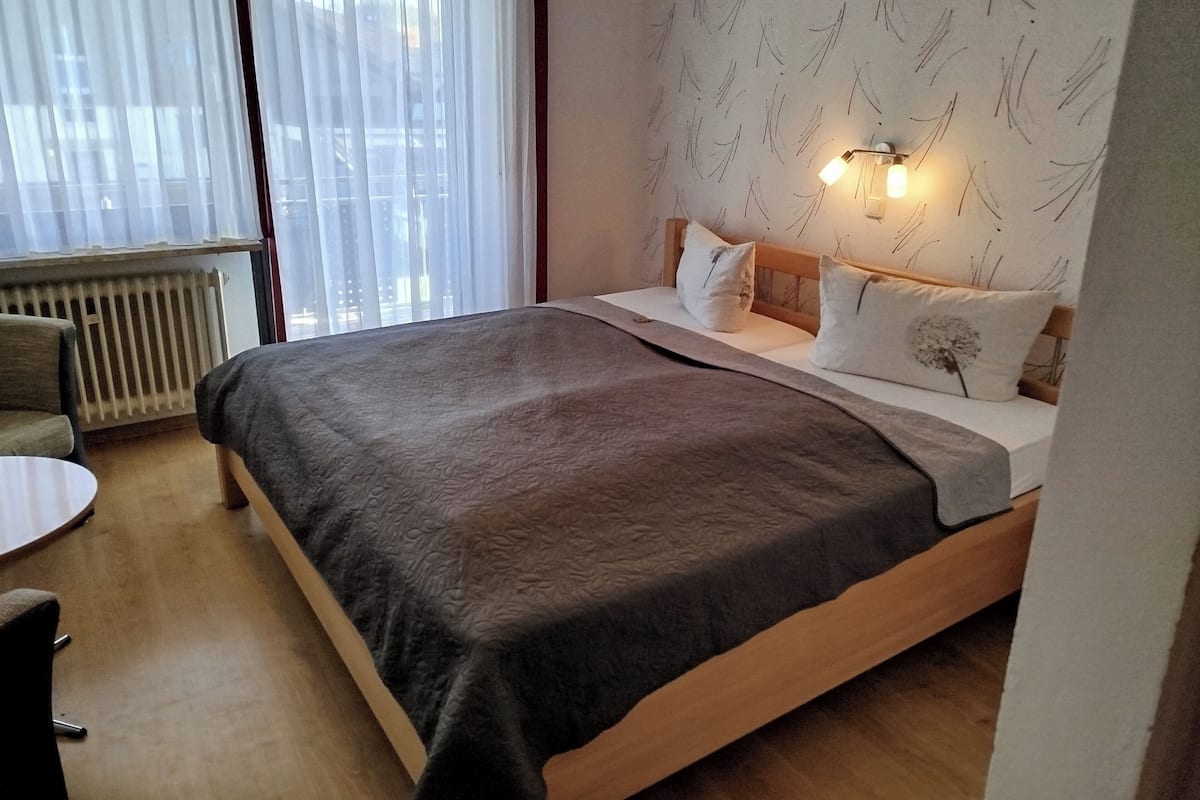 Ein Schlafzimmer mit Bett, Sessel, Heizkörper und einem Fenster mit Vorhängen.