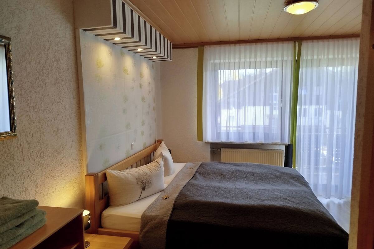 Ein Schlafzimmer mit Bett, Nachttisch, Kommode, einem Fenster mit durchscheinenden Vorhängen und einer Wandleuchte.