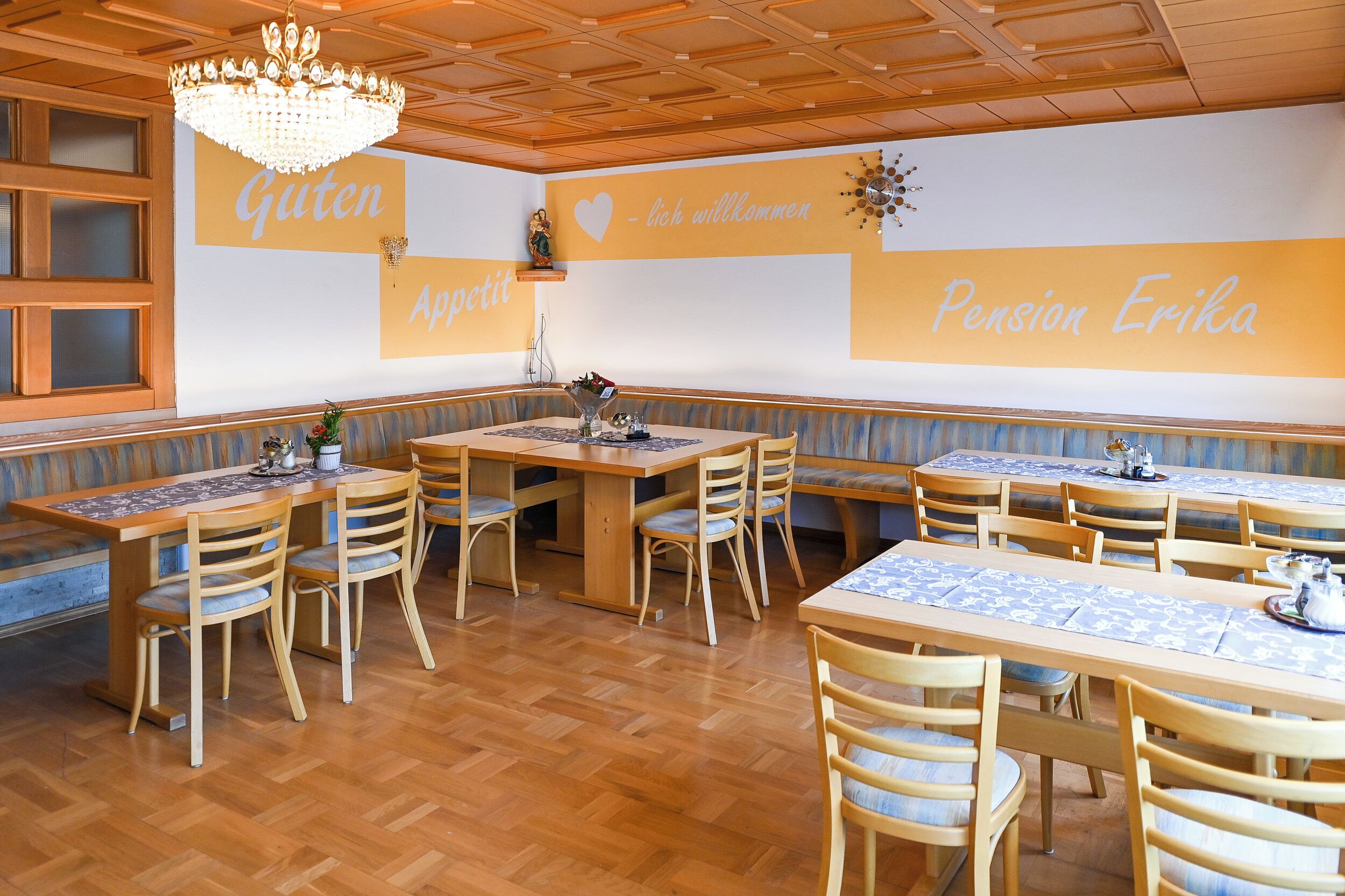 Un restaurante con mesas y sillas de madera, una araña y una pared con texto en alemán.
