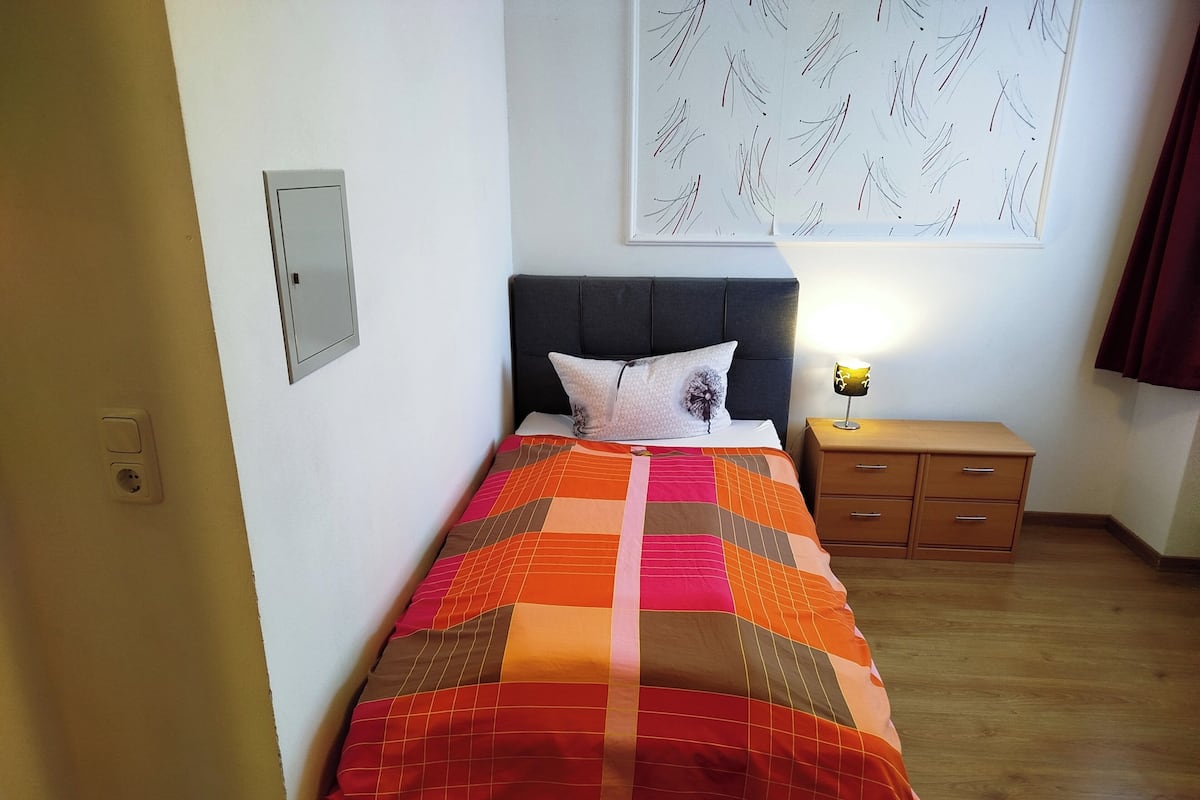 Ein ordentlich bezogenes Bett mit einer farbenfrohen Steude, ein Nachttisch mit einer Lampe und eine Wand mit einem gemusterten Kunstwerk.
