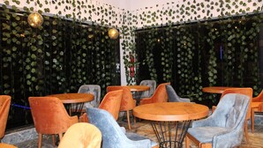 Interior de restaurante com mesas e cadeiras redondas, teto com luminárias pendentes e paredes cobertas por folhagem verde.