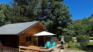 A wooden cabin with a porch, outdoor seating, and a stone fireplace. - Cocooning riverside Chalet on a small campsite in the Heart of Auvergne (Saint-Bonnet-de-Condat)