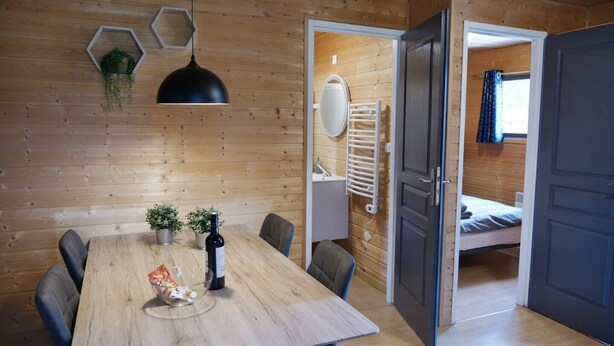 A wooden interior with a dining table, chairs, a hanging light fixture, a bed, and a door leading to another room. - Cocooning riverside Chalet on a small campsite in the Heart of Auvergne (Saint-Bonnet-de-Condat)
