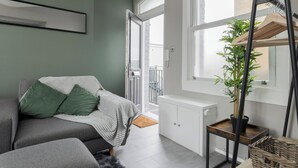 Moderni olohuone, jossa on harmaa sohva, pieni pöytä, jossa on ruukkukasvi, ja lasiovesta avautuva näkymä parvekkeelle.