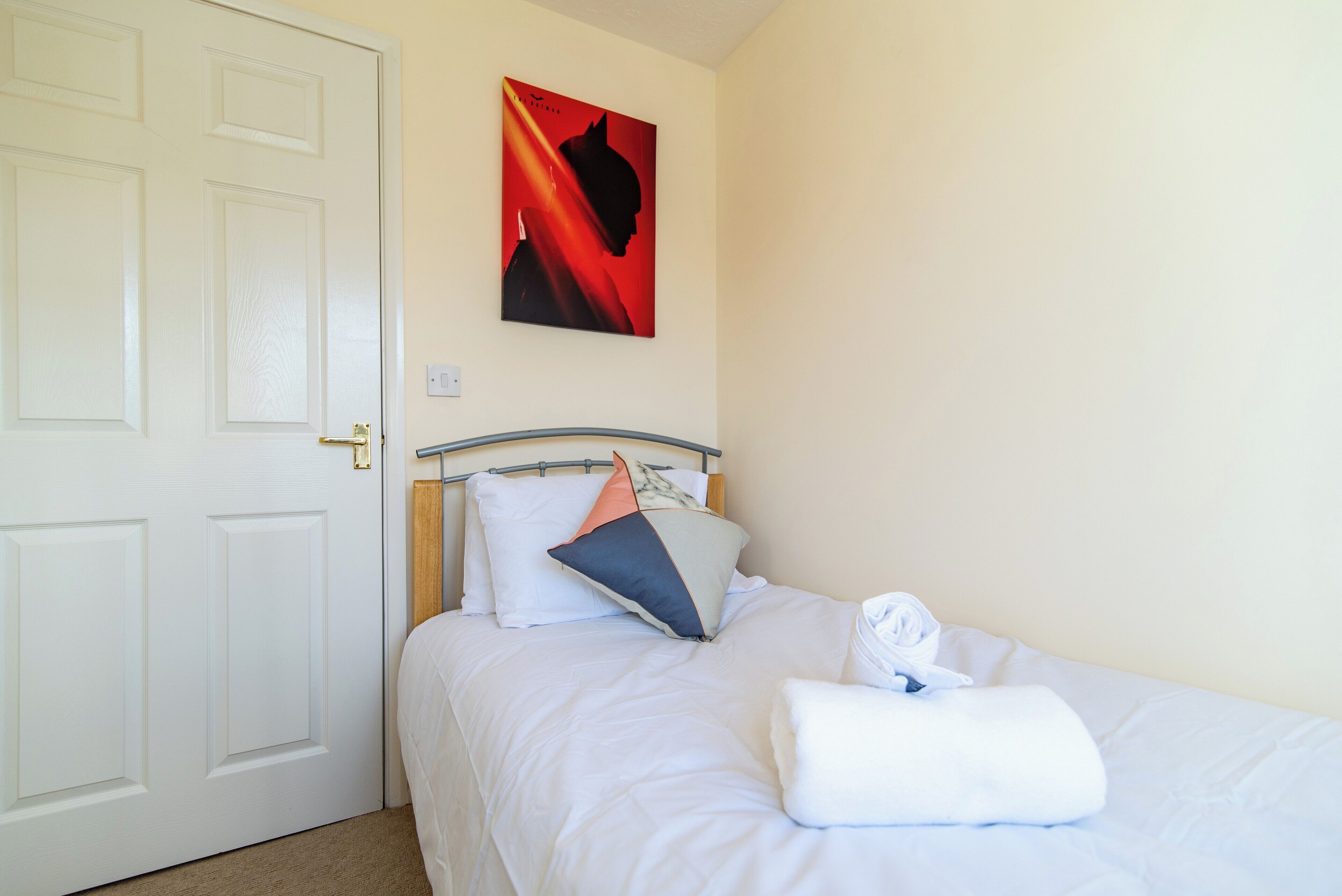 Ein ordentlich bezogenes Bett mit weißer Bettwäsche und einem gefalteten Handtuch, ein an der Wand hängendes Bild und eine geschlossene Tür.