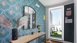 Ein Zimmer mit ornamentalem Wandpapier, einem Holzkonsolentisch, einem Spiegel, einer Tür mit Blick auf eine Stadtlandschaft und einer Fußmatte.