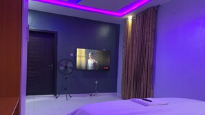 Egy modern hálószoba, melyben egy nagy ágy, egy falra szerelt, síkképernyős televízió és egy mennyezeti ventilátor található.
