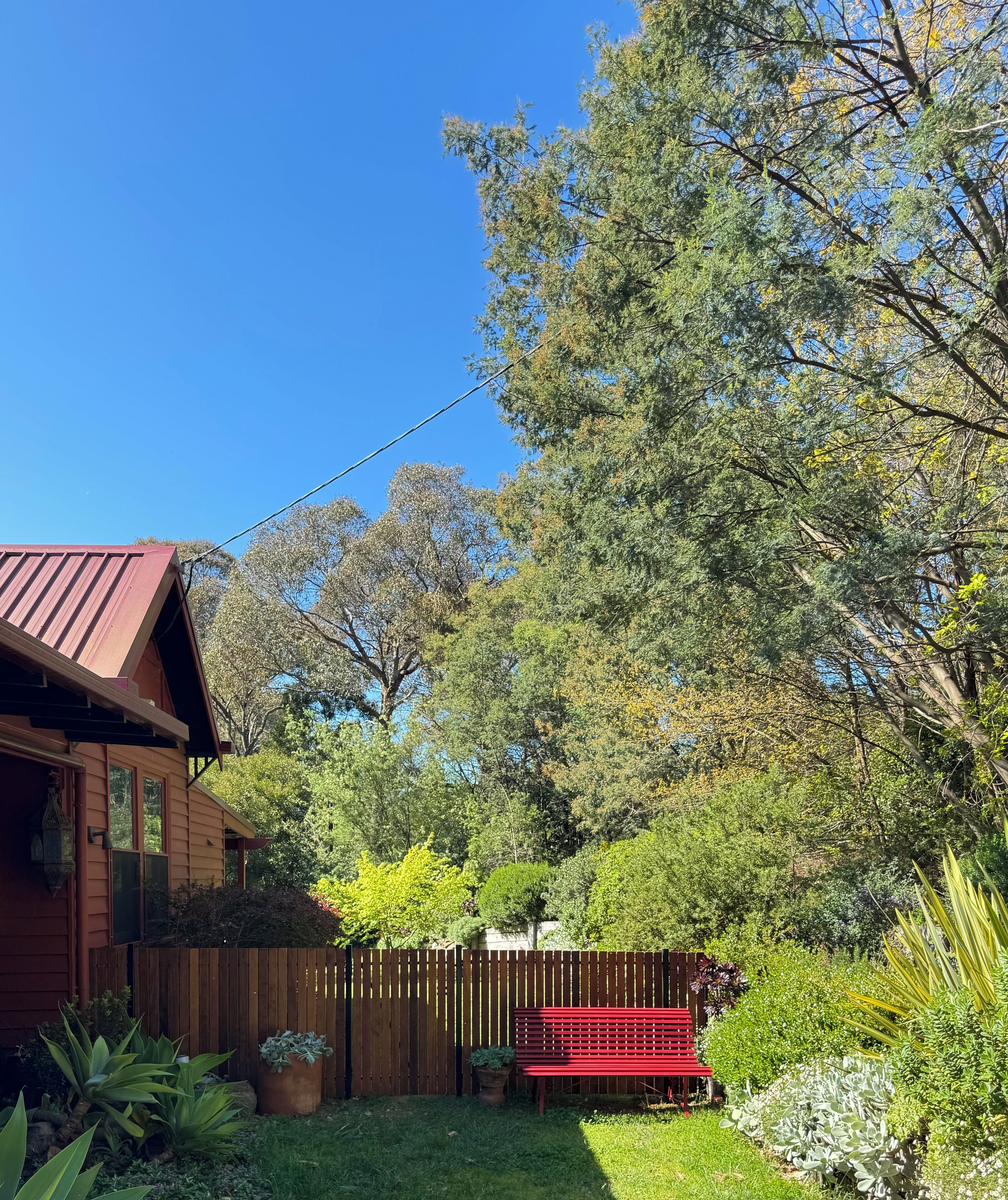 A wooden house with a red roof, surrounded by a wooden fence and lush greenery.