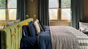 Une chambre à coucher avec une tête de lit recouverte d’un tissu vert, un grand lit avec une couvre-lit matelassé, deux lampes de chevet et une vue sur la verdure par les fenêtres.