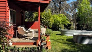Une maison en bois rouge avec un porche couvert, une chaise blanche et un petit jardin agrémenté de plantes en pot et d’une pelouse.