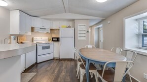 A compact kitchen with white cabinets, a microwave, a stove, and a refrigerator. There is a wooden dining table with chairs, and a window with blinds.