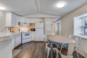 A compact kitchen with white cabinets, a microwave, a stove, and a refrigerator. There is a wooden dining table with chairs, and a window with blinds.