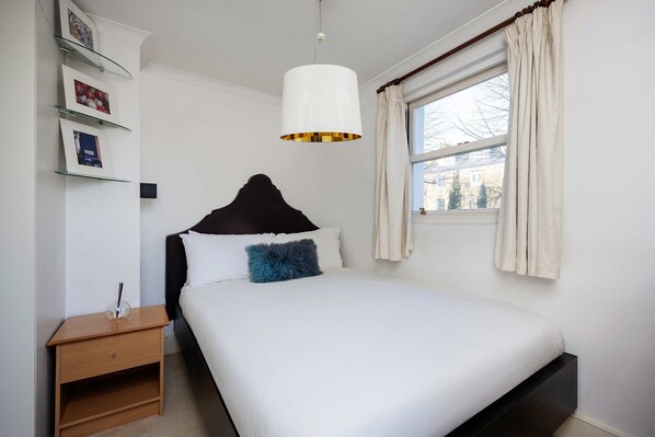 Ein ordentlich eingerichtetes Schlafzimmer mit Bett, Nachttisch und einem Fenster mit Vorhängen.