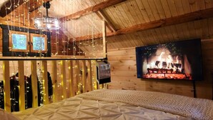 Una habitación acogedora con cama, un televisor mostrando una chimenea y luces colgantes.