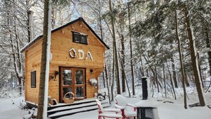 Una cabaña de madera con letrero "ODA", chimenea, un porche con sillas rojas y un bosque nevado de fondo.