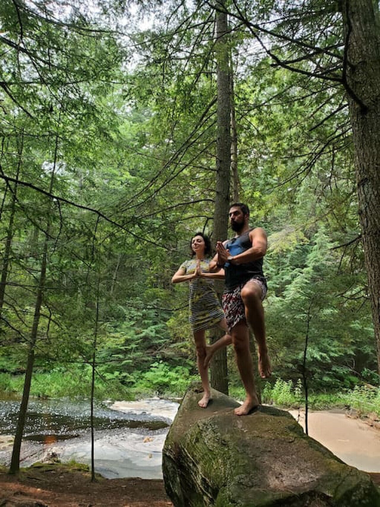 Two people standing on a rock in a forested area with a river in the background.