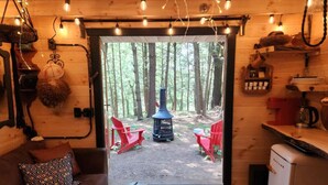 Un interior acogedor de cabaña con detalles de madera, un sofá, dos sillones rojos, una pequeña cocina americana y vistas a un bosque a través de un ventanal.
