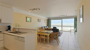 Una cocina con lavavajillas, un comedor con mesa y sillas, y vista a la playa a través de amplias ventanas.