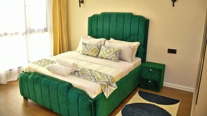 A green upholstered bed with patterned pillows, a bedside table, and a nightstand. - Sky park view apartment (Syokimau)