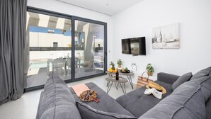 Ein modernes Wohnzimmer mit grauen Sofas, einem Flachbildfernseher, einem Essbereich mit Tisch und Stühlen sowie ein Balkon mit Pool.