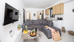 Eine moderne Wohnung mit grauem Ecksofa, einem flachbildfernseher an der Wand, einer kleinen Küche mit Holzschränken und einem Esstisch mit Früchten und einer Flasche.