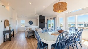 A modern dining area with a glass table and blue chairs, a dark kitchen in the background, and a view of the landscape through large windows. - Blue Moon - 4x4 Carova Beach, Ocean View, Pool, Hot Tub, Rec Room, Pool Table (4x4 - Carova NC)