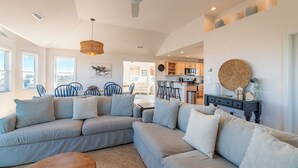 A spacious living area with a large L-shaped sectional sofa, a dining table with chairs, a kitchen area, and a ceiling fan. - Blue Moon - 4x4 Carova Beach, Ocean View, Pool, Hot Tub, Rec Room, Pool Table (4x4 - Carova NC)