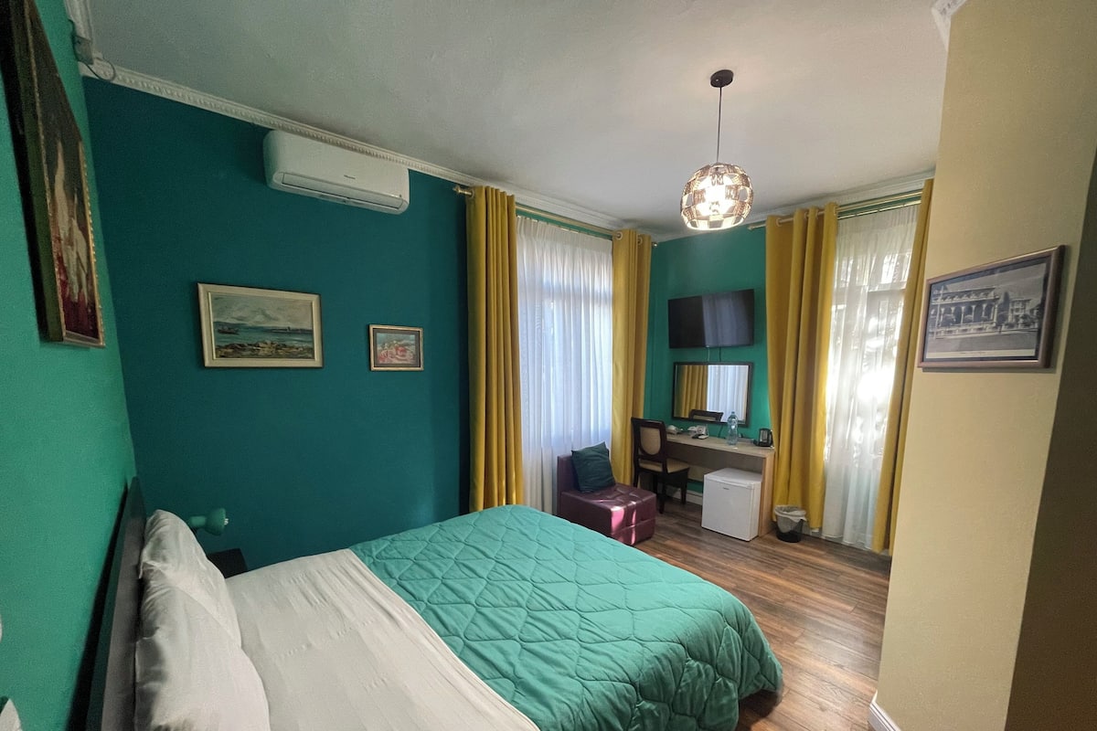 Une chambre aux murs de couleur turquoise, un lit recouvert d’une couette blanche et verte, un bureau avec une chaise, un petit réfrigérateur et un téléviseur fixé au mur.
